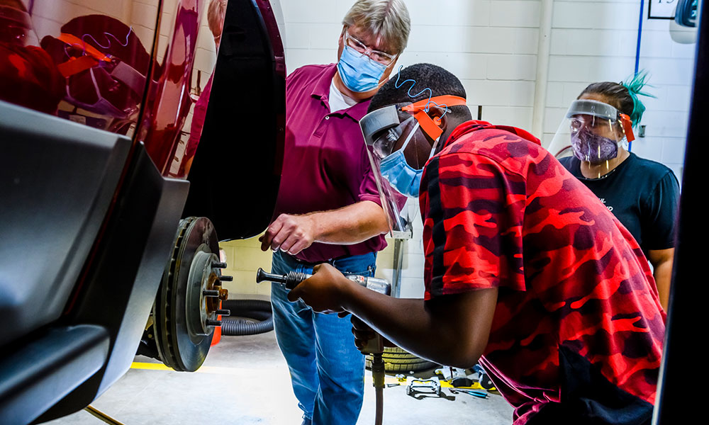 man working on a cars wheel axle while instructor and student look on behind him