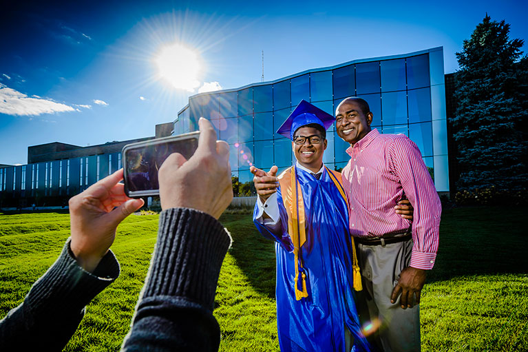 a father and his son, dressed in graduation cap and gown, posing for a photo outside the gannon building on the day of commencement ceremonies