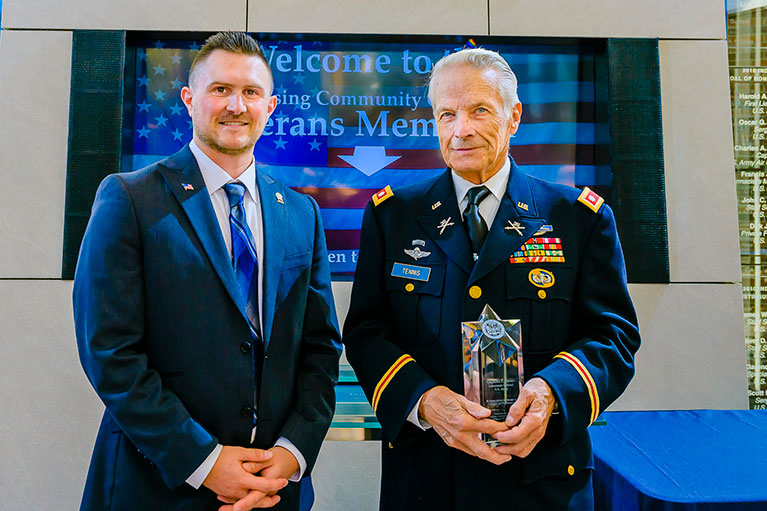 an older and younger veteran, posing for the camera at the veterans memorial center while the elder is holding an award he's received