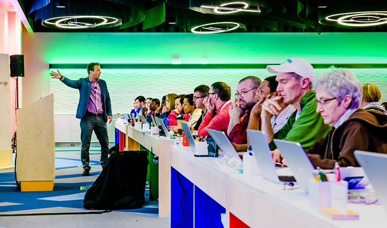 a room full of people looking laptops with a speaker pointing at a projector screen