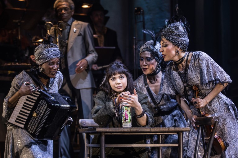 Several women in 1920's stage garb look over a seated woman from the musical Hadestown