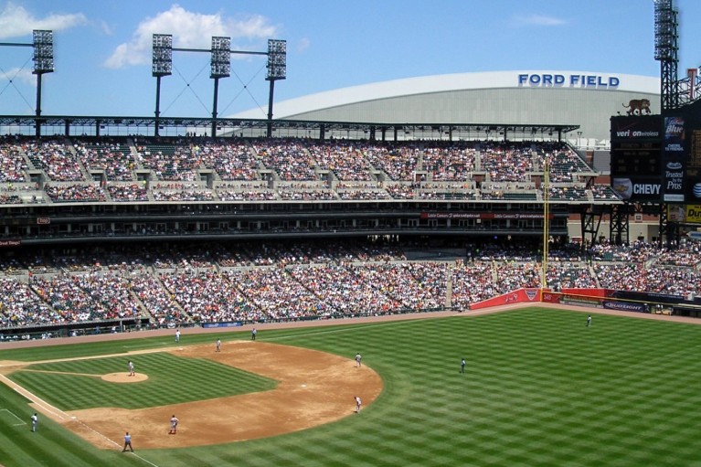 A baseball diamond with players in the foreground with fans in the stands on a blue sky day at Comerica Park