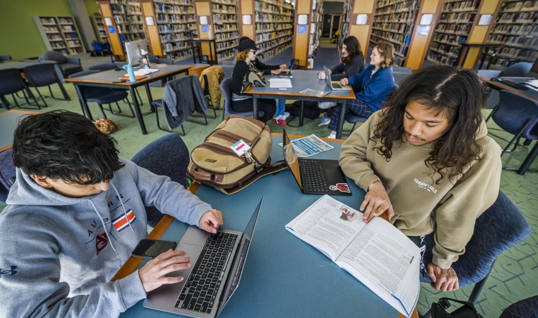Students sitting at library tables with technology and books spread out.