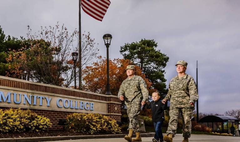 A man and a woman in milatary fatigues holding hands with a child in front of a Lansing Community College sign.