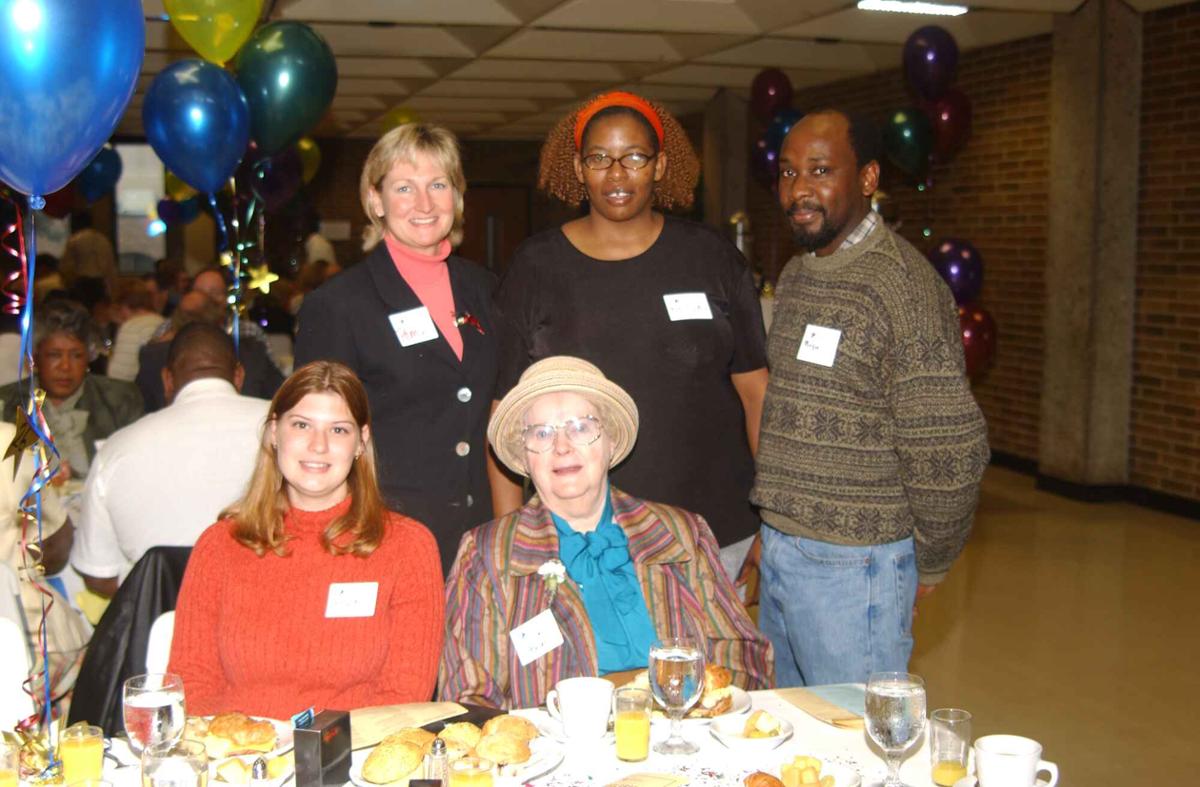 Two people sit at a table with food on it with three people standing behind them. There are balloons on the tables and around the room.