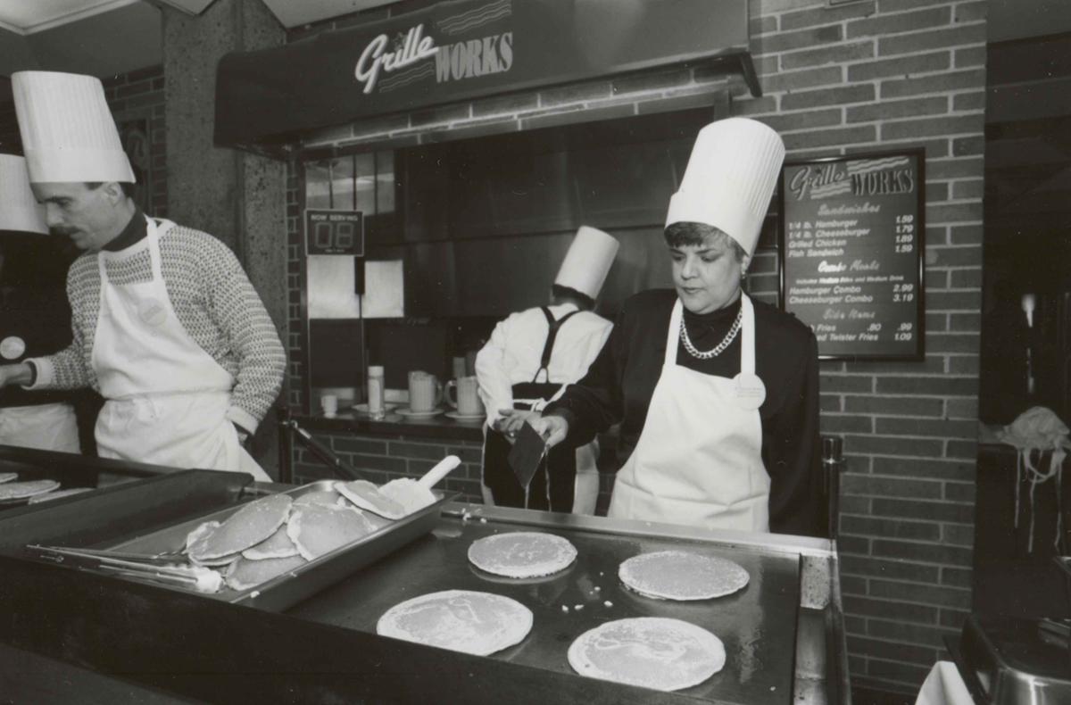 A man and a woman make pancakes at a griddle.