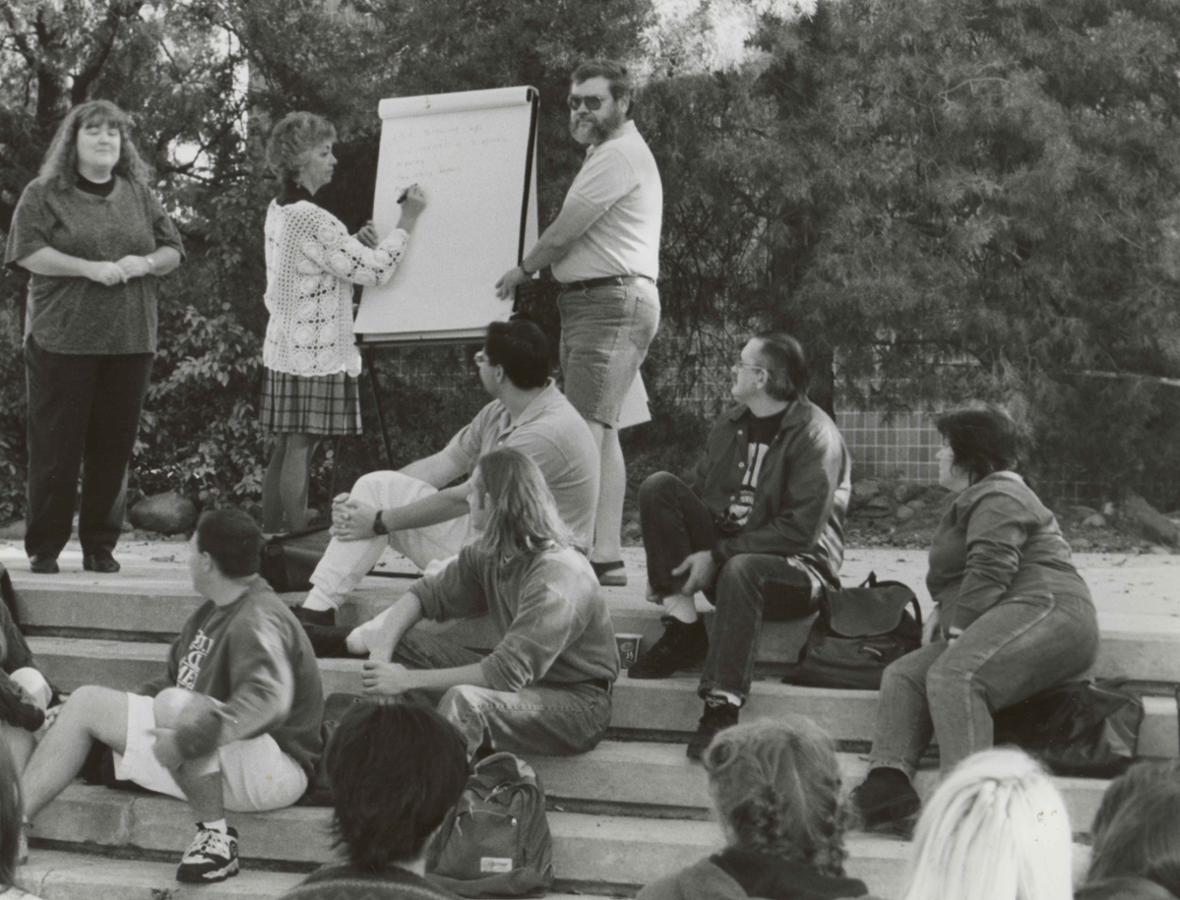 Outside, a woman writes on a large notepad on an easel, while a man stands on the other side of the notepad. A group of people look on.