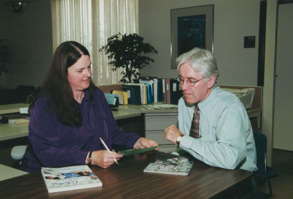 A woman and man sit at a table in an office looking at a piece of paper in the woman's hand.