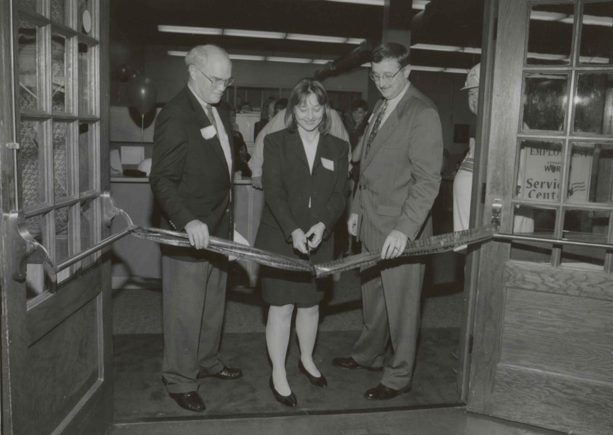 A woman cuts a ribbon strung across a doorway. A man stands on either side of her.