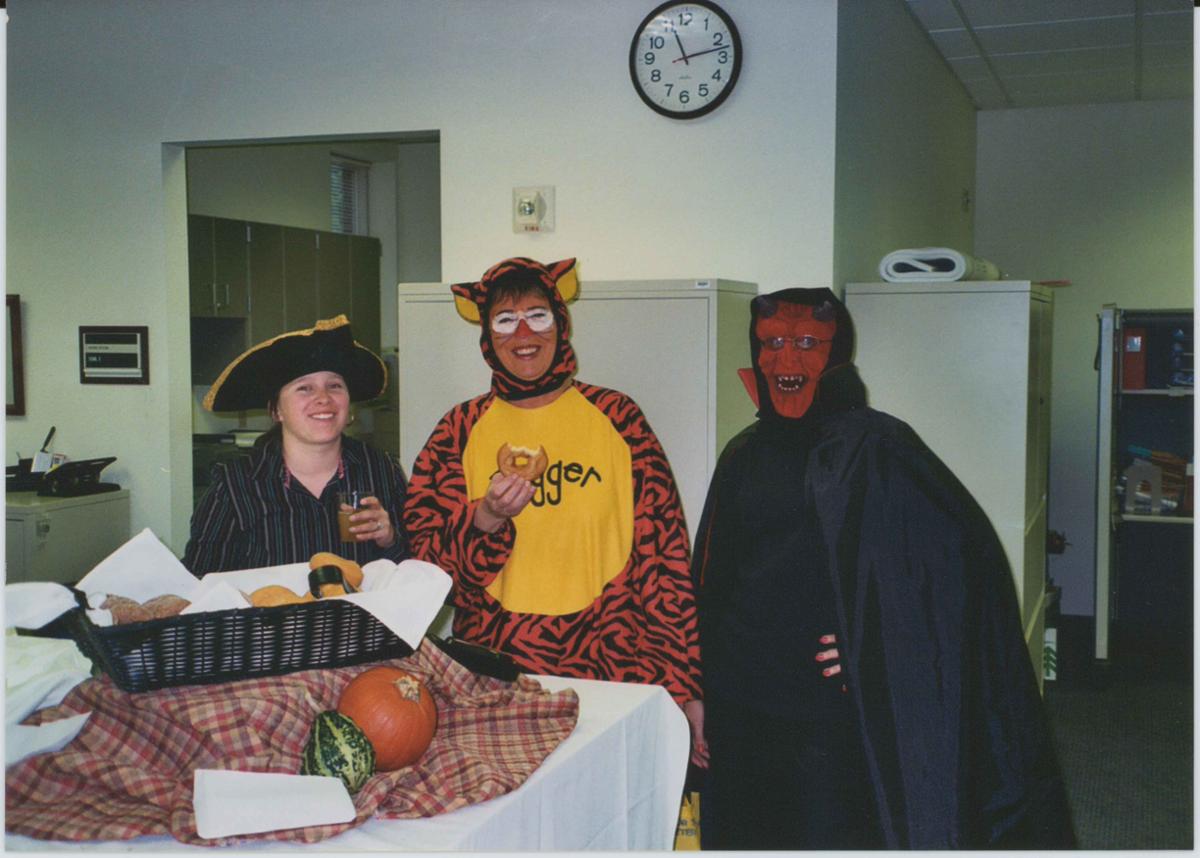 Three people stand in an office. The person on the left has on a pirate hat and is holding a drink. The person in the center is dressed up as Tigger and is holding a donut with a bite taken out of it. The person on the left is dressed in black with a cape and has on a red mask with red lines on the face, horns, and sharp teeth.
