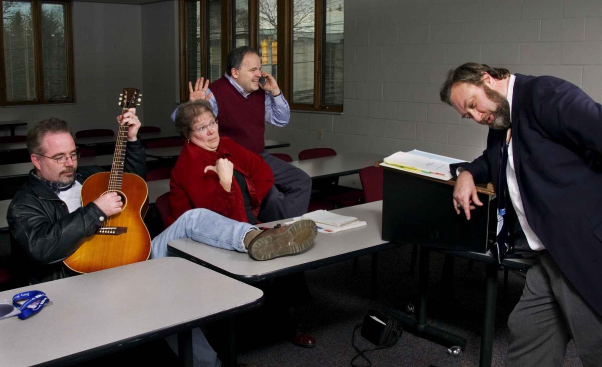 Four people in a classroom. The man on the left sits with his leg on the table playing guitar. The woman next to him points at him with her thumb. The man behind her sits on the table and talks on the phone. A man stands at the front of the classroom with his arm on the lectern and his head facing down.