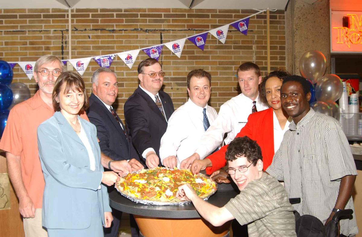 A group of people holds up a dish of nachos at a table. A Taco Bell bunting hangs on a wall behind them.