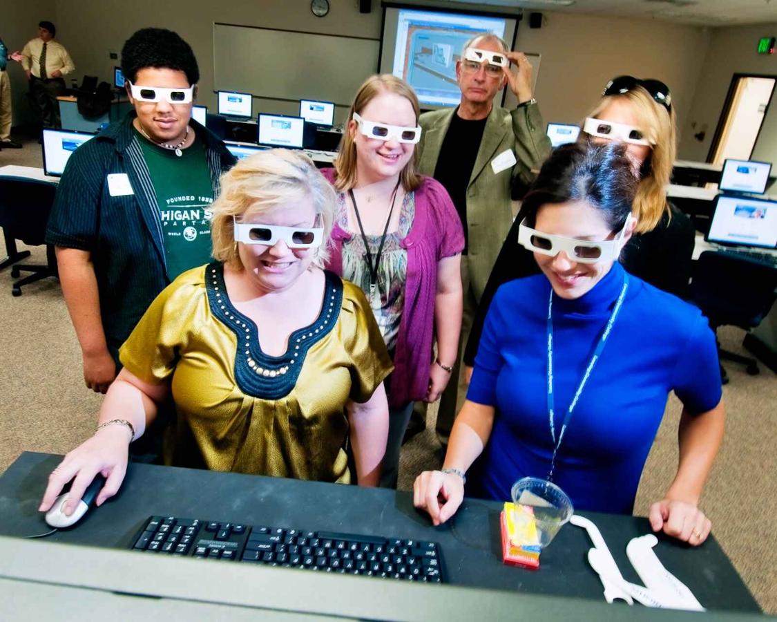 A group of people wear 3D glasses and look at a screen in a computer lab.
