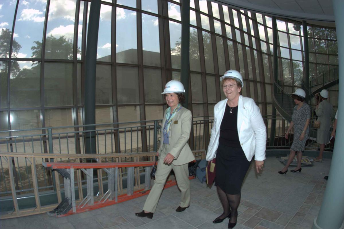 Two women wearing hard hats walk in front of windows with ladders lying on the ground next to them.
