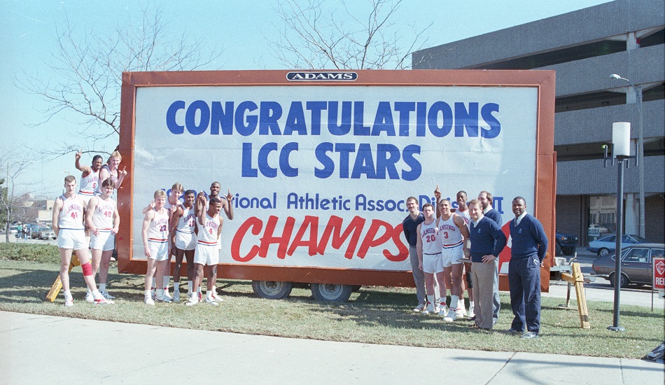 The 1988 Men's Basketball team and coaching staff stand by the "Congratulations LCC Stars, 1988 National Athletic Assoc. Division II Champs!" billboard at the corner of Shiawassee Street and Capitol Avenue. (from left): Pat Archer, Mark Watts, Kirk Simon, Trent Jakus, Jeff Casler, Carter Briggs, Ed Banks, Kirk Baker, Quinn Ferguson, Assistant Coach Steve Schmitt, Todd Eaton, Marcell Cole, Melvin Kelly, Head Coach Art Frank, Assistant Coach Paul Stoll, Assistant Coach Mike Ingram - March 1988