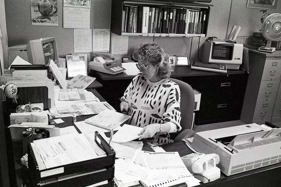 An employee works on paper documents in an office containing some of the early technology used on campus - ca. 1980s