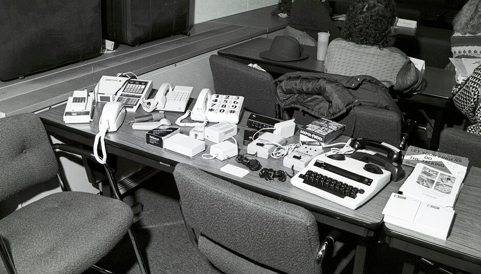 Telephone equipment displayed on a table in a classroom includes early telecommunications devices used by the hearing impaired, such as a teletypewriter (bottom right in photo) - ca. 1980s